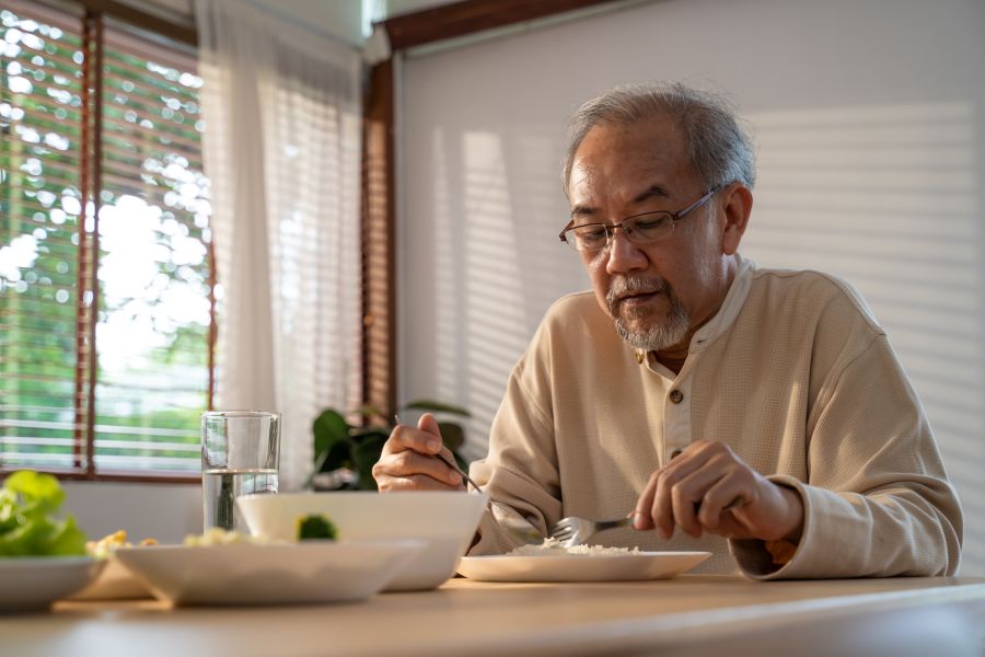 Unhappy Asian Senior older man sit alone, eat foods on table in house. Depressed mature attractive elderly retired grandfather stay at home with painful face feeling upset, lonely and miss his family